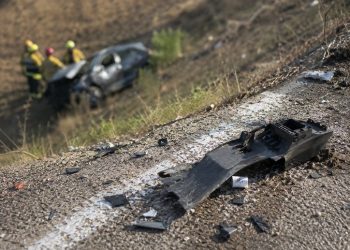 Close-up of crash debris near a freeway embankment in daylight, with a blurred damaged vehicle and emergency responders visible farther below the roadway.