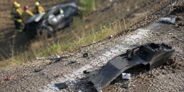 Close-up of crash debris near a freeway embankment in daylight, with a blurred damaged vehicle and emergency responders visible farther below the roadway.