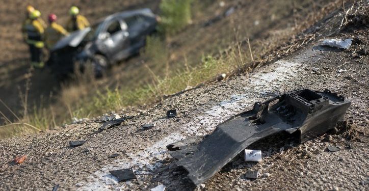 Close-up of crash debris near a freeway embankment in daylight, with a blurred damaged vehicle and emergency responders visible farther below the roadway.