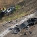 Close-up of crash debris near a freeway embankment in daylight, with a blurred damaged vehicle and emergency responders visible farther below the roadway.