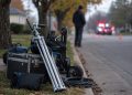 Close-up of film production equipment along a residential curb, with a blurred crew member standing near distant police lights in the background.