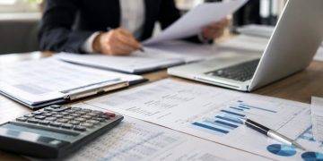 Close-up of financial documents, a calculator and a laptop on an office desk, with a blurred employee reviewing records in the background.