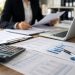 Close-up of financial documents, a calculator and a laptop on an office desk, with a blurred employee reviewing records in the background.