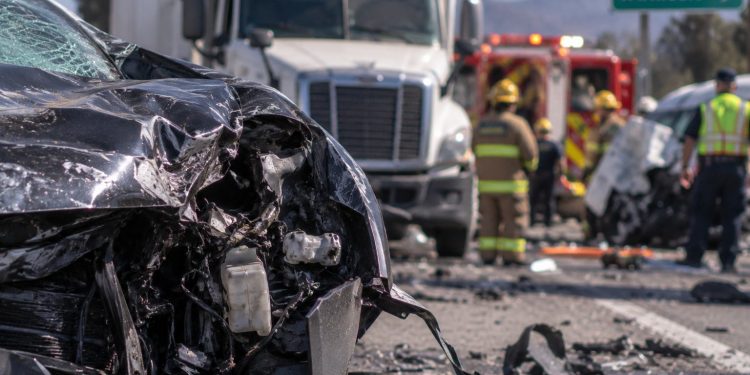 Close-up of heavily damaged vehicles at a multi-vehicle crash scene, with a big rig, emergency crews and a blocked Southern California roadway blurred in the background.