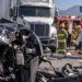 Close-up of heavily damaged vehicles at a multi-vehicle crash scene, with a big rig, emergency crews and a blocked Southern California roadway blurred in the background.