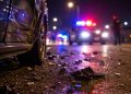 Close-up of roadway debris beside a damaged vehicle at night, with blurred police lights and an officer standing in the distance.