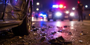 Close-up of roadway debris beside a damaged vehicle at night, with blurred police lights and an officer standing in the distance.