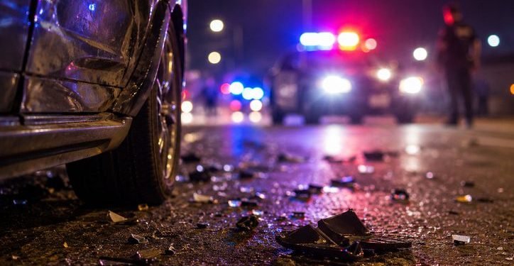 Close-up of roadway debris beside a damaged vehicle at night, with blurred police lights and an officer standing in the distance.