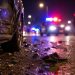Close-up of roadway debris beside a damaged vehicle at night, with blurred police lights and an officer standing in the distance.