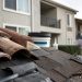 Close-up of roof tiles and repair materials beside a condominium building, with a blurred resident reviewing paperwork in the background.