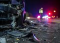 Close-up of scattered crash debris beside a damaged vehicle at night, with a blurred officer and emergency lights visible farther down the roadway.