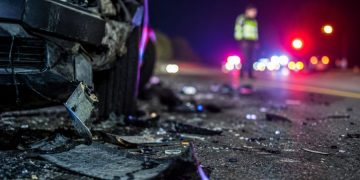 Close-up of scattered crash debris beside a damaged vehicle at night, with a blurred officer and emergency lights visible farther down the roadway.