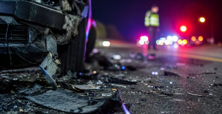 Close-up of scattered crash debris beside a damaged vehicle at night, with a blurred officer and emergency lights visible farther down the roadway.