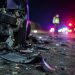 Close-up of scattered crash debris beside a damaged vehicle at night, with a blurred officer and emergency lights visible farther down the roadway.