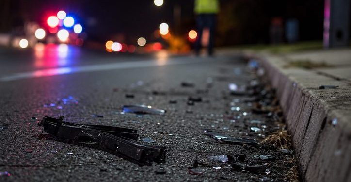 Close-up of scattered roadway debris near the edge of a dark street, with a blurred responder standing beside distant emergency lights.