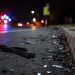 Close-up of scattered roadway debris near the edge of a dark street, with a blurred responder standing beside distant emergency lights.