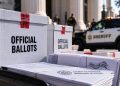 Close-up of sealed election ballot boxes and official election documents near a courthouse, with a blurred sheriff's vehicle in the background, illustrating a ballot custody case.