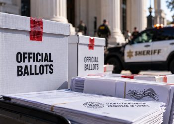 Close-up of sealed election ballot boxes and official election documents near a courthouse, with a blurred sheriff's vehicle in the background, illustrating a ballot custody case.