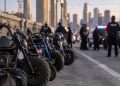 Close-up of several minibikes lined up on a downtown Los Angeles bridge with blurred police vehicles and officers in the background.