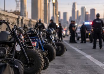 Close-up of several minibikes lined up on a downtown Los Angeles bridge with blurred police vehicles and officers in the background.