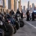 Close-up of several minibikes lined up on a downtown Los Angeles bridge with blurred police vehicles and officers in the background.