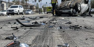 Close-up of shattered automotive debris and skid marks on a sunlit roadway, with an overturned vehicle and a blurred responder visible in the background.