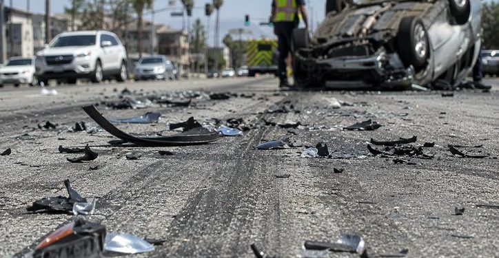 Close-up of shattered automotive debris and skid marks on a sunlit roadway, with an overturned vehicle and a blurred responder visible in the background.