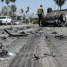 Close-up of shattered automotive debris and skid marks on a sunlit roadway, with an overturned vehicle and a blurred responder visible in the background.