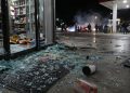 Close-up of shattered glass and scattered store merchandise near a gas station entrance, with blurred figures and tire smoke visible farther outside.