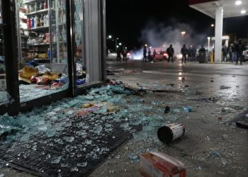 Close-up of shattered glass and scattered store merchandise near a gas station entrance, with blurred figures and tire smoke visible farther outside.