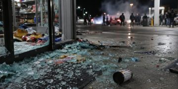 Close-up of shattered glass and scattered store merchandise near a gas station entrance, with blurred figures and tire smoke visible farther outside.