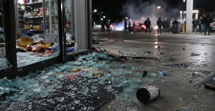 Close-up of shattered glass and scattered store merchandise near a gas station entrance, with blurred figures and tire smoke visible farther outside.