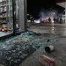 Close-up of shattered glass and scattered store merchandise near a gas station entrance, with blurred figures and tire smoke visible farther outside.