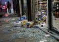Close-up of shattered storefront glass and scattered grocery items near an entrance, with a blurred person standing outside and emergency lights visible in the background.