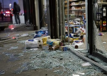 Close-up of shattered storefront glass and scattered grocery items near an entrance, with a blurred person standing outside and emergency lights visible in the background.