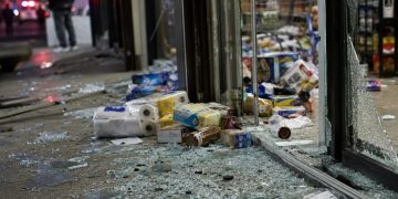 Close-up of shattered storefront glass and scattered grocery items near an entrance, with a blurred person standing outside and emergency lights visible in the background.