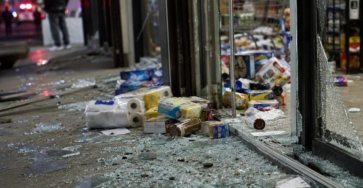 Close-up of shattered storefront glass and scattered grocery items near an entrance, with a blurred person standing outside and emergency lights visible in the background.