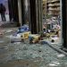 Close-up of shattered storefront glass and scattered grocery items near an entrance, with a blurred person standing outside and emergency lights visible in the background.