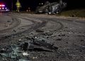 Close-up of shattered vehicle debris and heavy tire marks along a dark roadway, with a blurred overturned car and emergency responder in the background.