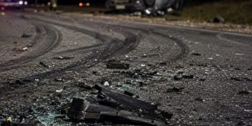 Close-up of shattered vehicle debris and heavy tire marks along a dark roadway, with a blurred overturned car and emergency responder in the background.