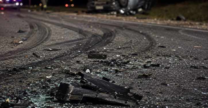 Close-up of shattered vehicle debris and heavy tire marks along a dark roadway, with a blurred overturned car and emergency responder in the background.