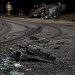Close-up of shattered vehicle debris and heavy tire marks along a dark roadway, with a blurred overturned car and emergency responder in the background.