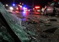 Close-up of shattered vehicle glass and damaged roadway debris beside blurred emergency lights at night, representing a serious multi-vehicle collision.