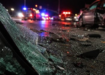 Close-up of shattered vehicle glass and damaged roadway debris beside blurred emergency lights at night, representing a serious multi-vehicle collision.