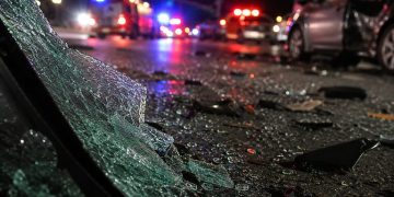 Close-up of shattered vehicle glass and damaged roadway debris beside blurred emergency lights at night, representing a serious multi-vehicle collision.