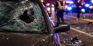 Close-up of shattered windshield glass and roadway debris beside a dark sedan, with blurred investigators and emergency lights in the background.