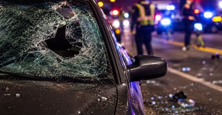 Close-up of shattered windshield glass and roadway debris beside a dark sedan, with blurred investigators and emergency lights in the background.