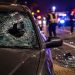 Close-up of shattered windshield glass and roadway debris beside a dark sedan, with blurred investigators and emergency lights in the background.