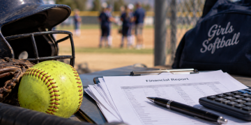 Close-up of softball equipment and financial records on a table, with a blurred youth baseball field in the background, illustrating an embezzlement investigation.