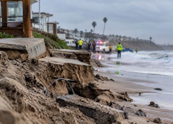 Close-up of storm-damaged coastal sand and erosion near a Southern California beach, with blurred emergency activity in the background.